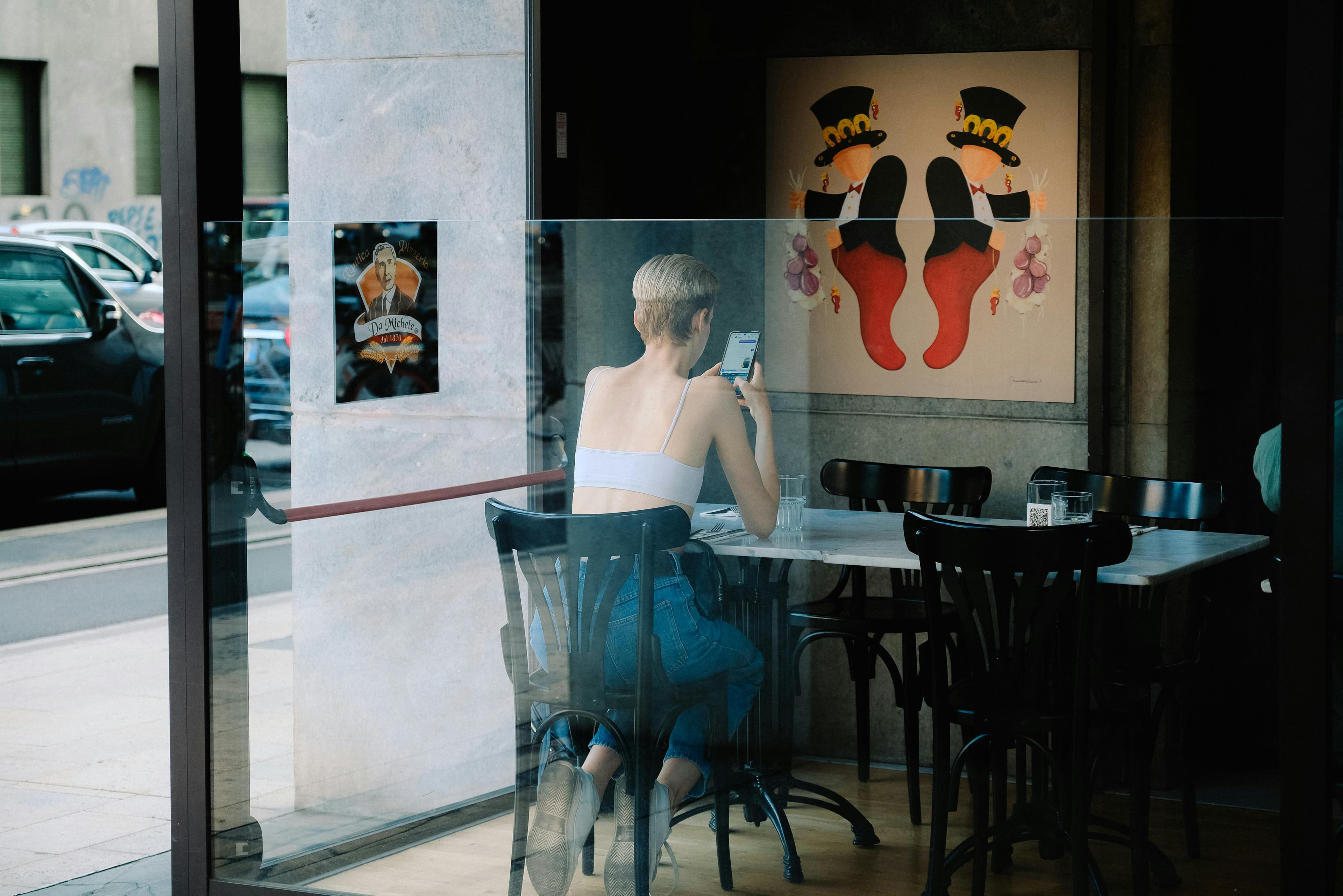 Person seated alone in a cafe, viewed through glass while checking a phone