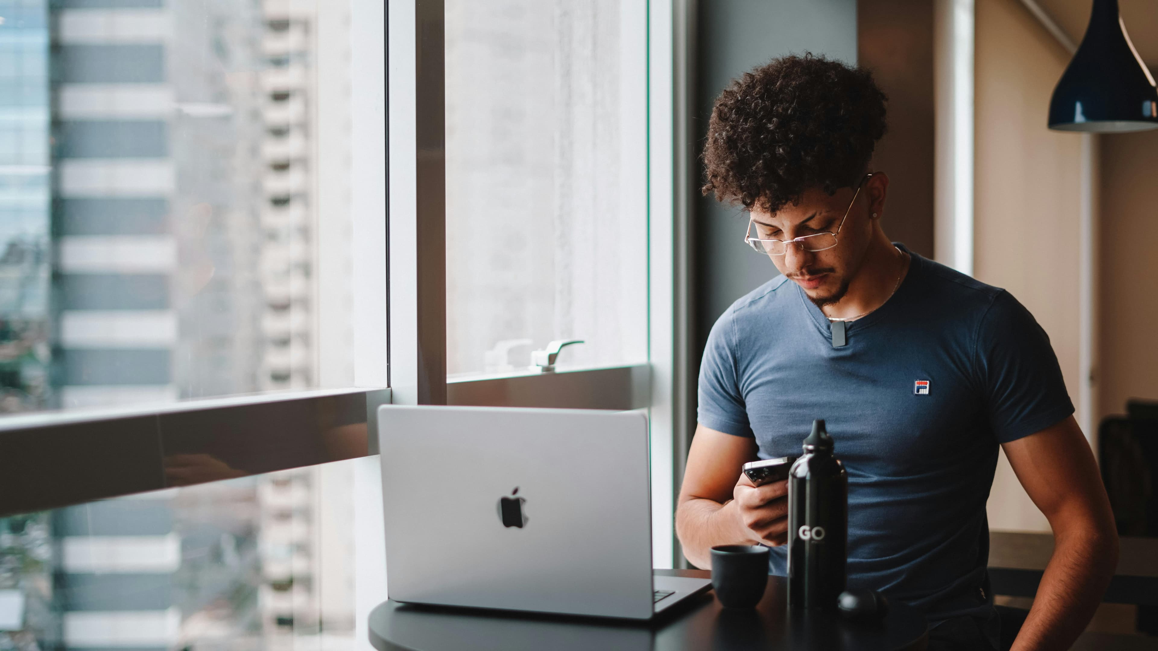 Young man checking his phone beside a laptop in a bright cafe workspace
